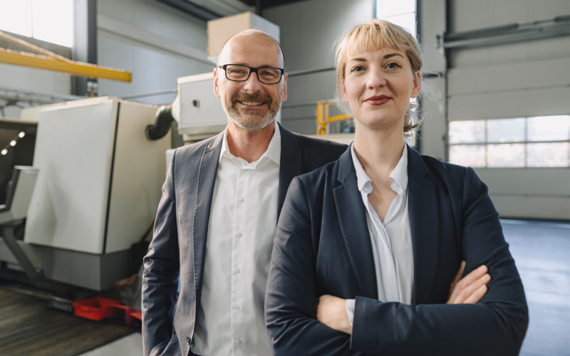 Eine Frau mit verschränkten Armen und ein Mann mit Hände in den Hosentaschen stehen vor Anlagen vor einem Rolltor