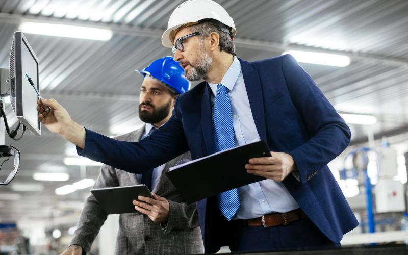 2 Männer mit Anzug und Helm in einer Produktionshalle schauen auf einen Bildschirm